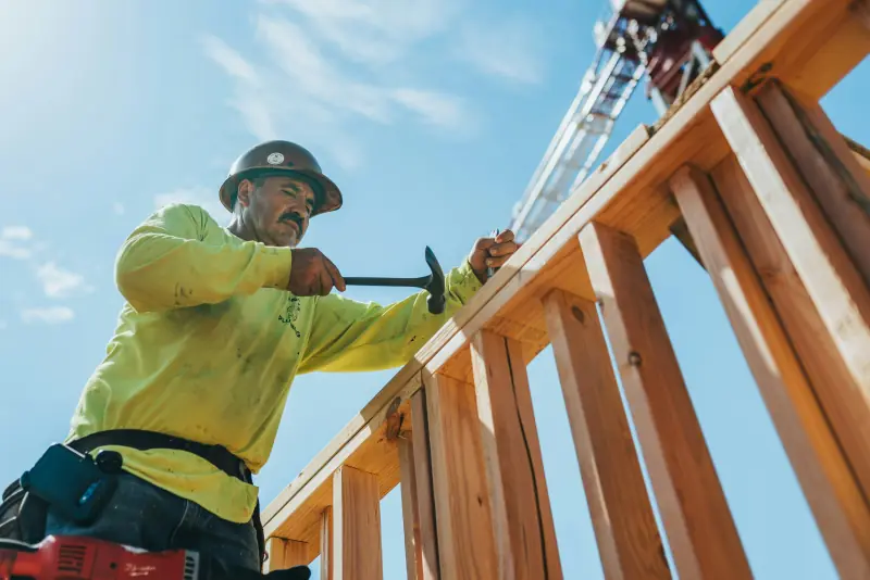 A carpenter in a hard hat nailing the top plate of a wood framed wall against a blue sky